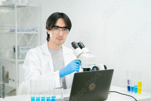 Fototapeta A scientist and researcher in laboratory setting is using microscope while wearing safety glasses and gloves, with test tubes and laptop nearby. scientist researcher chemistry laboratory