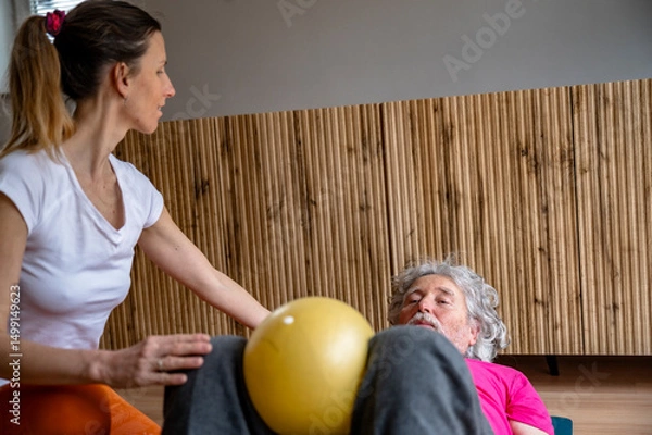 Obraz Senior man exercising with physiotherapist using a fitness ball indoors