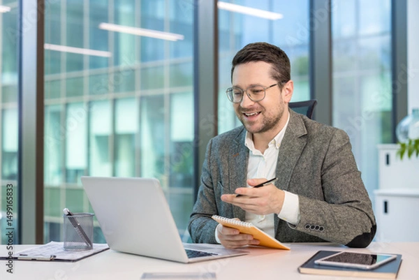 Obraz Smiling young businessman in a suit sitting at his workplace in the office and talking remotely via laptop, making notes in a notebook