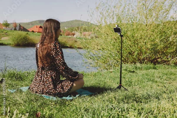 Fototapeta Young woman meditating by the river online. The concept of online tutorials, distance learning.