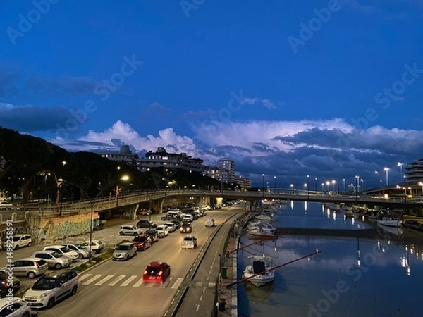 Fototapeta Twilight view of the Lungofiume dei Poeti in Pescara, Italy, with cars driving and boats moored in the canal