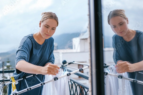 Obraz Young woman drying laundry on balcony during slow morning at home. Real moment, Gen Z lifestyle, sustainability, calm domestic routine, mindful living, eco-conscious habits, natural light.
