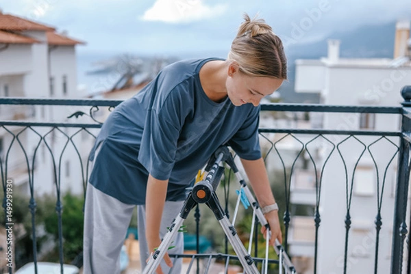 Obraz Young woman drying laundry on balcony during slow morning at home. Real moment, Gen Z lifestyle, sustainability, calm domestic routine, mindful living, eco-conscious habits, natural light.