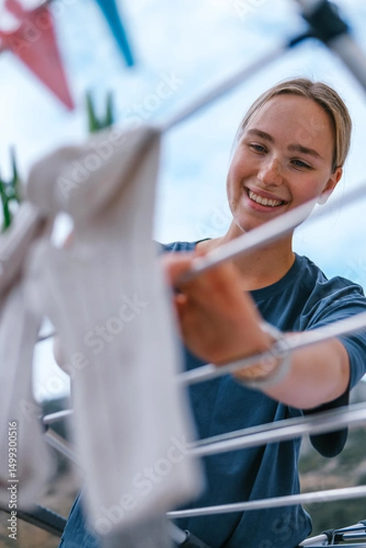 Obraz Young woman drying laundry on balcony during slow morning at home. Real moment, Gen Z lifestyle, sustainability, calm domestic routine, mindful living, eco-conscious habits, natural light.