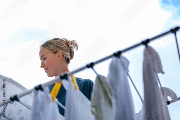 Obraz Young woman drying laundry on balcony during slow morning at home. Real moment, Gen Z lifestyle, sustainability, calm domestic routine, mindful living, eco-conscious habits, natural light.