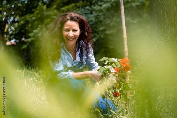 Obraz smiling middle-aged woman with red hair showing a bunch of flowers sitting in the grass