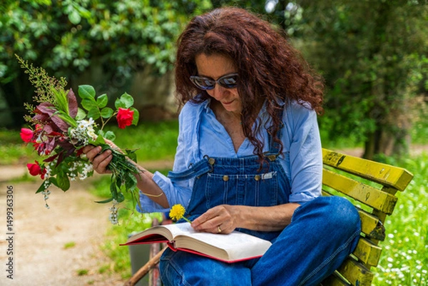 Obraz middle-aged woman with red hair sitting on a bench reading a book holding a bouquet of flowers