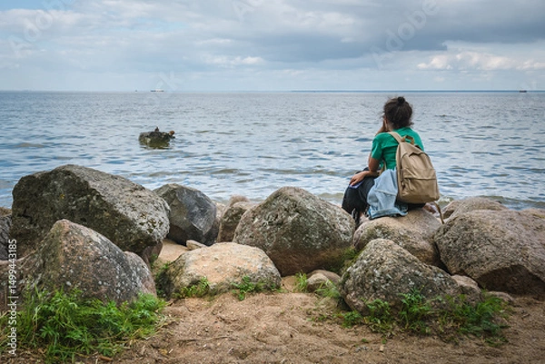 Fototapeta A girl tourist with a backpack sits on the rocks near the seashore with her back to us and looks into the distance.