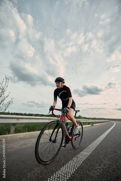 Fototapeta Confident girl, cyclist in full gear riding along open countryside road, powerful stride and focused eyes locked on route ahead. Concept of commitment, sport, movement, fitness journey.