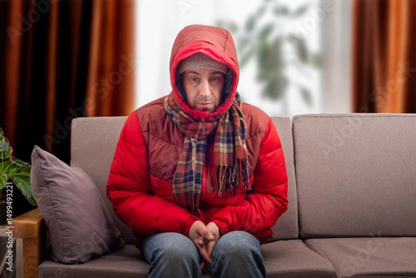 Obraz Middle age male model in red parka and checkered scarf getting cold sitting on the sofa in cold living room