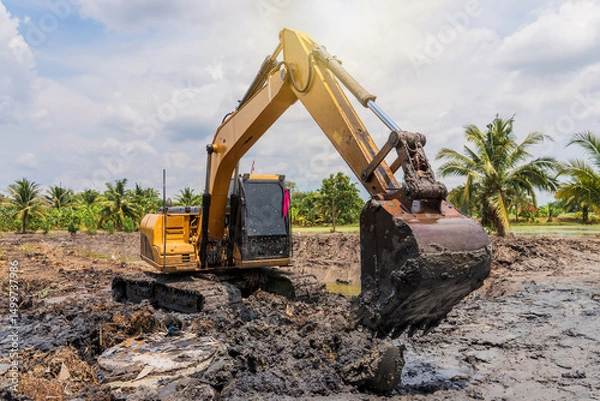 Fototapeta Excavator digging mud clay soil from pond bottom to expand and lift trench with level area for cultivation of garden furrows with water surrounding them in lowlands Thailand.