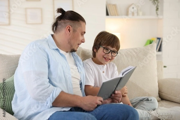Fototapeta Father and his son reading book together on sofa at home
