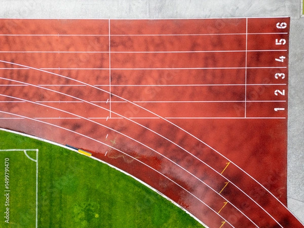 Fototapeta Aerial view of a red running track with marked lanes and curved lines. Athletic stadium section with part of a green football field. Sports and fitness concept.