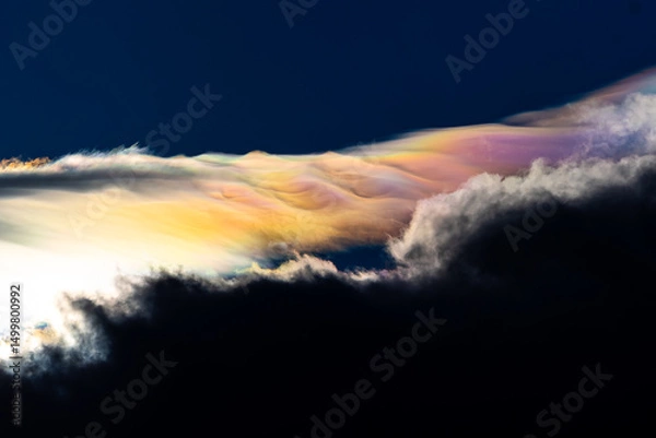 Fototapeta Spectacular iridescent cloud glows in vivid rainbow colors above dark storm clouds. Rare optical weather phenomenon captured against a deep blue sky. Amazing type of clouds.