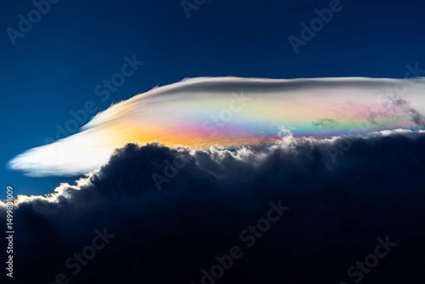 Fototapeta Spectacular iridescent cloud glows in vivid rainbow colors above dark storm clouds. Rare optical weather phenomenon captured against a deep blue sky. Amazing type of clouds.