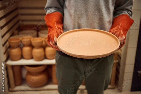 Fototapeta Ceramist standing with a clay plate by a kiln in his workshop