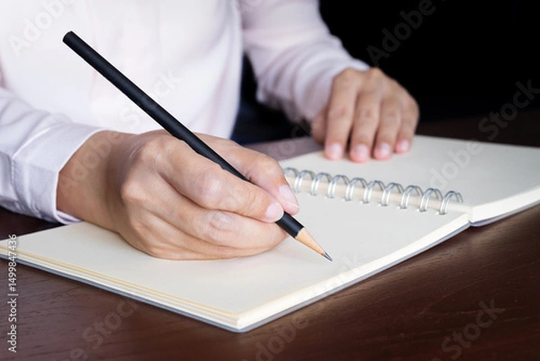 Fototapeta closeup view of woman hand using pencil writing on blank white page notebook while sitting at the table.