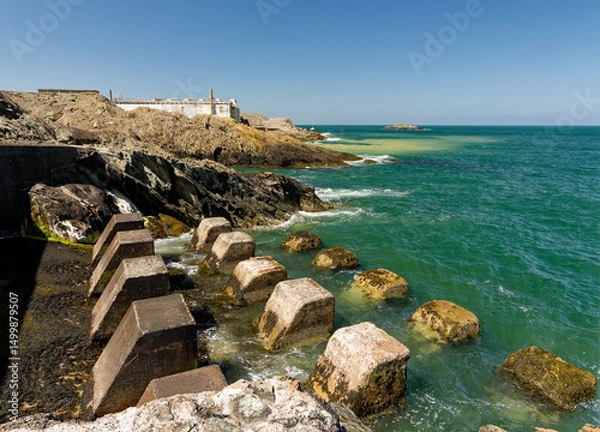 Fototapeta Scenic coastal view with rocky shoreline and concrete sea defenses vibrant turquoise waters under clear skies.