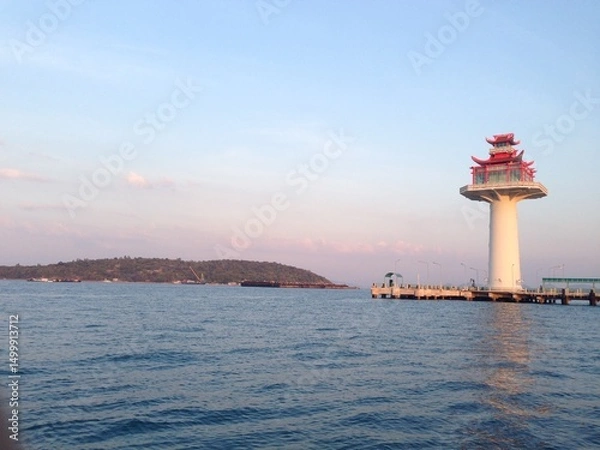 Fototapeta Scenic view of a lighthouse with traditional Asian architectural style on a pier at sunset, overlooking a calm sea and distant island. Peaceful maritime landscape perfect for travel or coastal themes.