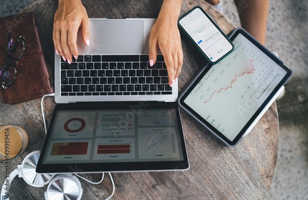 Fototapeta Top view of freelance workstation with woman typing on laptop, smartphone and tablet showing financial charts, illustrating multi-device productivity and tech-driven remote work