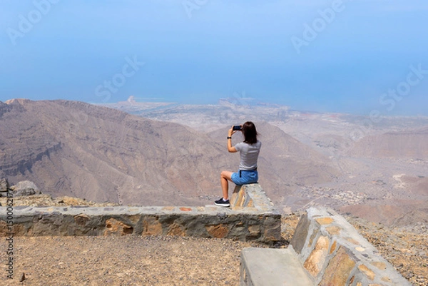 Fototapeta  Woman sitting on a stone barrier taking a photo of breathtaking mountain and sea view from Jebel Jais, the highest peak in the United Arab Emirates