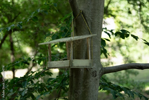 Fototapeta A wooden birdhouse hangs from a sturdy tree branch, surrounded by vibrant green leaves. Spring sunlight filters through the forest, creating a serene atmosphere