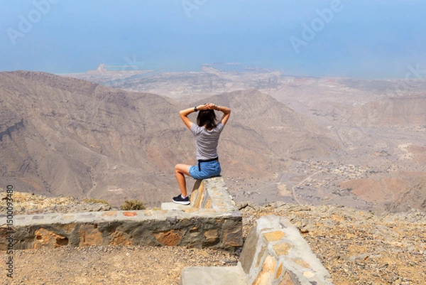 Fototapeta Woman sitting on a stone wall at the top of Jebel Jais, enjoying the vast mountain view and coastline below. Scenic, serene, breathtaking moment in Ras Al Khaimah mountains, UAE