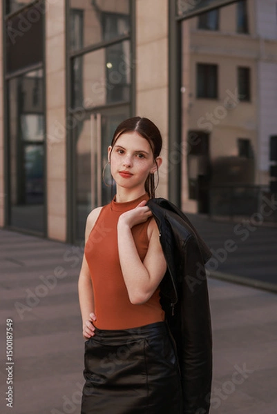 Fototapeta A girl with freckles in a leather skirt near a skyscraper.