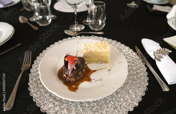 Fototapeta Gourmet gastronomy in a social event. Closeup view of a grilled loin fillet with sauce and a potato mille feuille in a white dish on the gala table with a beautiful tableware setting