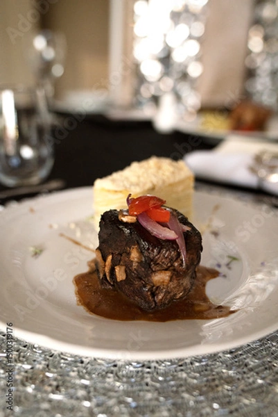 Fototapeta Gourmet gastronomy in a social event. Closeup view of a grilled loin fillet with sauce and a potato mille feuille in a white dish on the gala table
