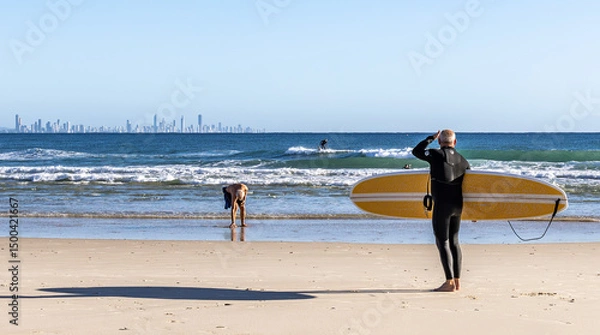 Fototapeta A surfer with board looking towards the water from Coolangatta Beach with the high-rise city skyscape of the Gold Coast on the horizon.