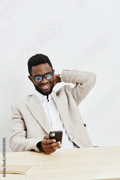 Fototapeta Smiling happy young man with afro hairstyle wearing glasses and beige blazer sitting at desk using smartphone, isolated on plain white background. Casual, cheerful, adult, elearning, technology