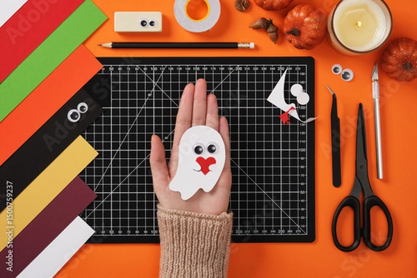 Obraz In the open palm of hand lies a finished paper craft of a ghost in love. There are paper crafting supplies around. Cutting mat, scissors, tweezers, cutter, pencil, eraser.