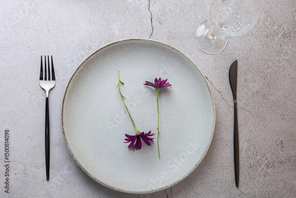 Obraz Top view of ceramic plate with two violet flowers, black cutlery and crystal glass on light grey background. Clean and tidy composition. Concept of elegant table and natural style