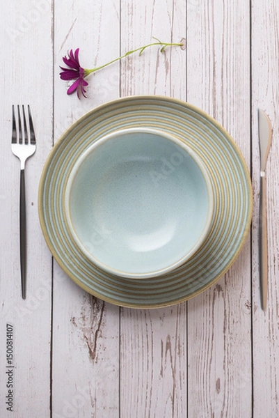 Obraz Top view of a light blue ceramic plate on a dinner plate with cutlery and a purple flower on light wooden background. Concept of elegant table setting, minimalism and natural design