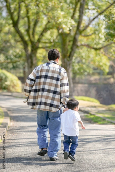 Fototapeta In March, at a sunny forested park in Japan, a 3-year-old Japanese boy walks hand in hand with his father. Captured from behind, this photo shows a quiet and heartwarming moment during a spring walk.
