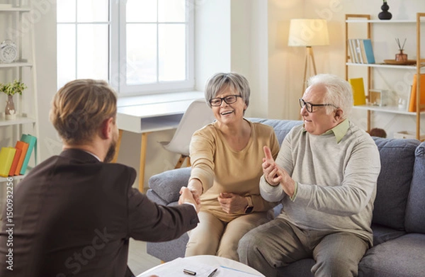 Fototapeta Portrait of happy cheerful senior couple sitting on couch in living room at home shaking hands with male financial advisor reaching agreement in health insurance. Investment in retirement concept