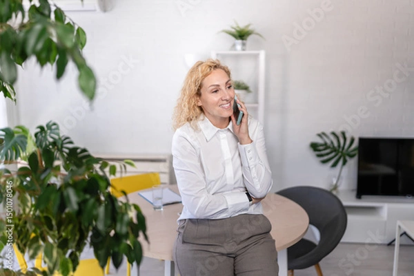 Fototapeta Smiling businesswoman talking on the phone in a modern office, standing confidently by her desk, engaged in a pleasant and professional conversation.