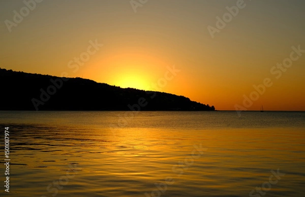 Fototapeta Tranquil seascape at sunset with golden reflections on the calm water and the sun setting behind a silhouette of a coastal hill.