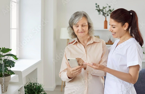 Fototapeta Young friendly smiling female nurse showing elderly woman how to use smartphone. Caregiver teaching senior person to handle with mobile phone. Nursing home service and patient support concept.