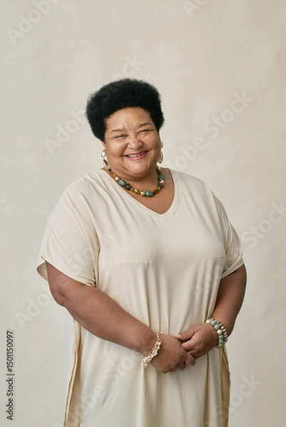 Fototapeta Vertical portrait of Black woman standing against neutral background with relaxed smile and hands clasped while wearing beaded necklace and bracelet posing in studio