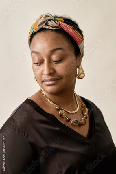 Fototapeta Vertical shot of Black woman wearing colorful headscarf and matching jewelry looking down with contemplative expression while posing in studio against beige background