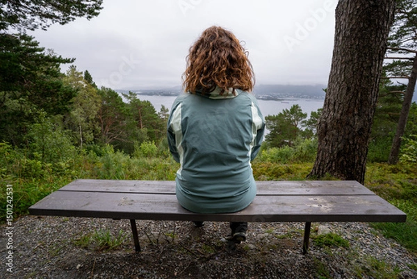 Fototapeta Curly-haired woman sitting alone on a bench, gazing at the Alesund fjord under a cloudy sky, surrounded by lush pine forest and peaceful nature