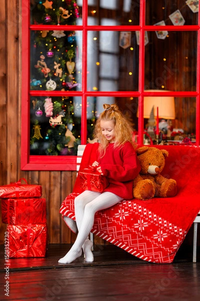 Obraz A little girl at home in a red sweater near a Christmas tree celebrates Christmas and New Year.
