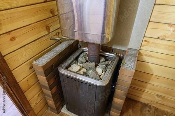 Fototapeta sauna stove filled with rocks sits in a corner of a wooden sauna. The stove has a metal chimney and brick surrounding. The wall is made of light-colored wood planks