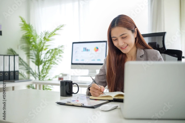 Obraz Professional businesswoman taking notes while working on laptop in modern office.