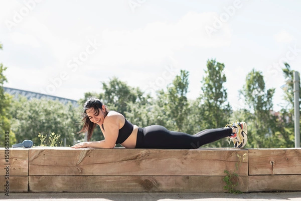 Fototapeta A young woman laughs happily as she relaxes between workouts, lying on a bench in the park