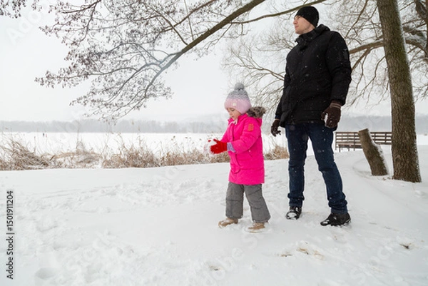 Fototapeta Father with daughter dressed warmly and in warm gloves builds a snowman in a snowy near the lake.