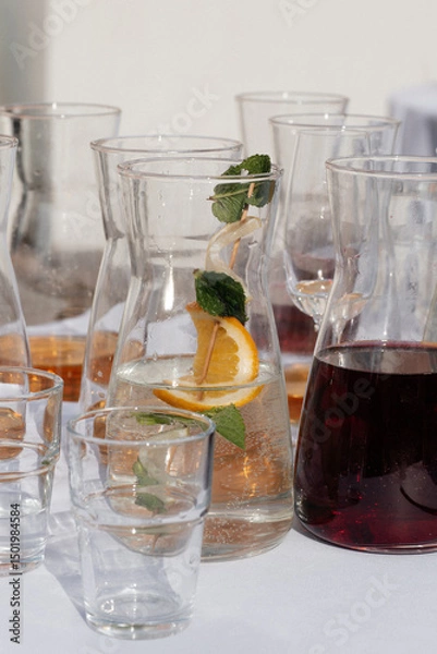 Obraz Glass jugs with water, lemon, mint and juice on a white table in sunlight. Refreshing summer drinks served outdoors, perfect for event, party or restaurant design use.
