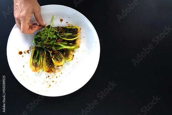 Obraz Hand adds finishing touches to a plate of bok choy with herbs in a stylish kitchen setting.